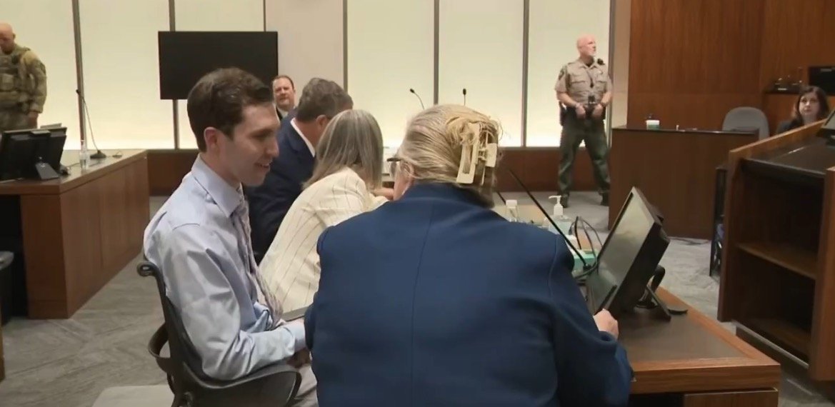 Courtroom scene featuring a group of individuals seated at a table, with law enforcement present in the background, during a legal proceeding.